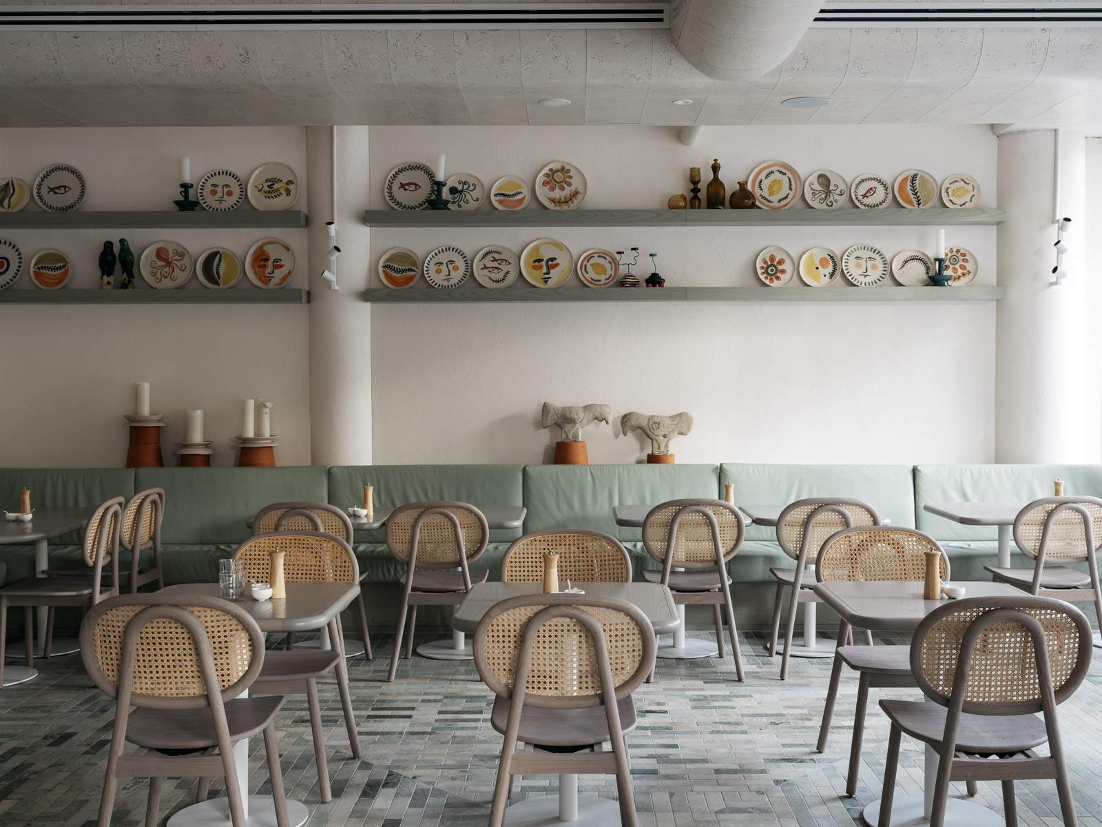 The interiors of a restaurant, with multiple tables, grey chairs, a light green banquette and white walls with ceramic on shelves