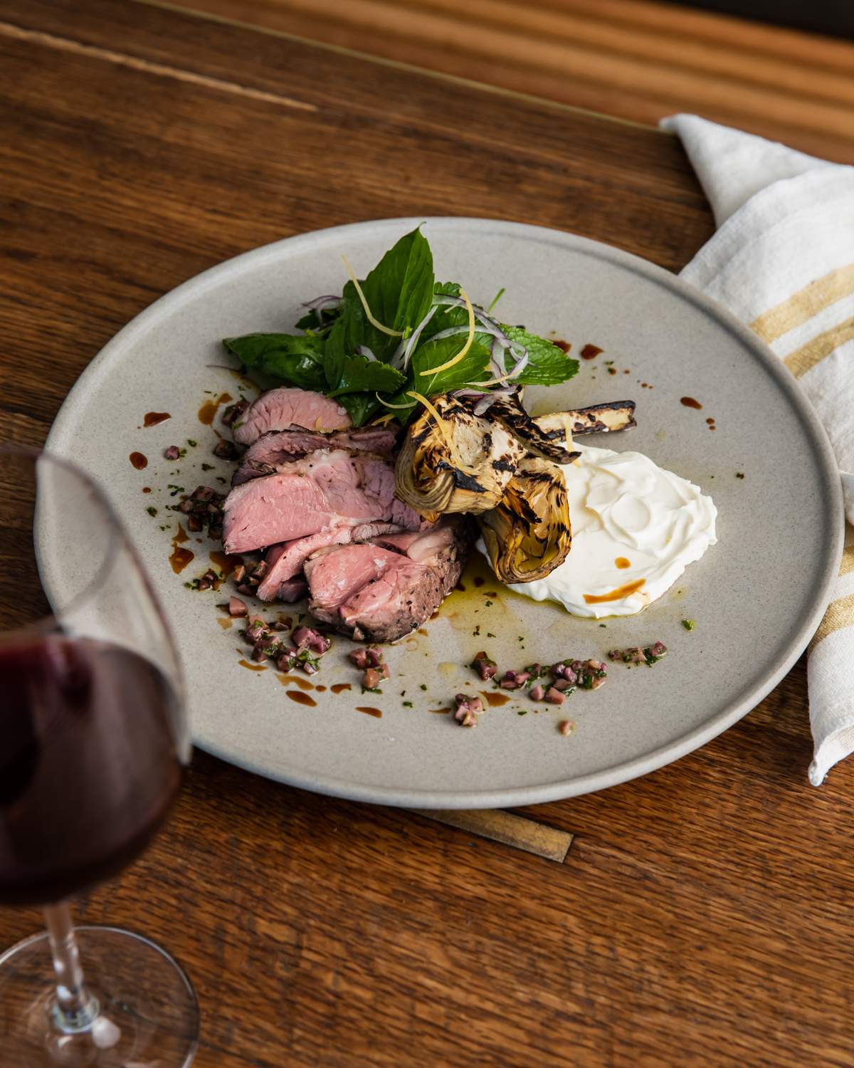 A grey plate with roast lamb rump, artichokes and labneh next to a glass of red wine