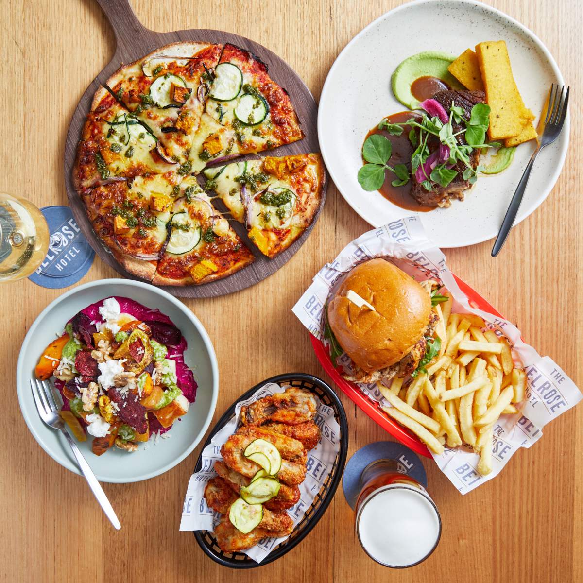 An array of dishes on a table that includes a vegetable pizza, a colourful salad bowl, chicken wings, a burger with chips and a beef brisket with polenta fires, with a pint of beer and a glass of white wine.