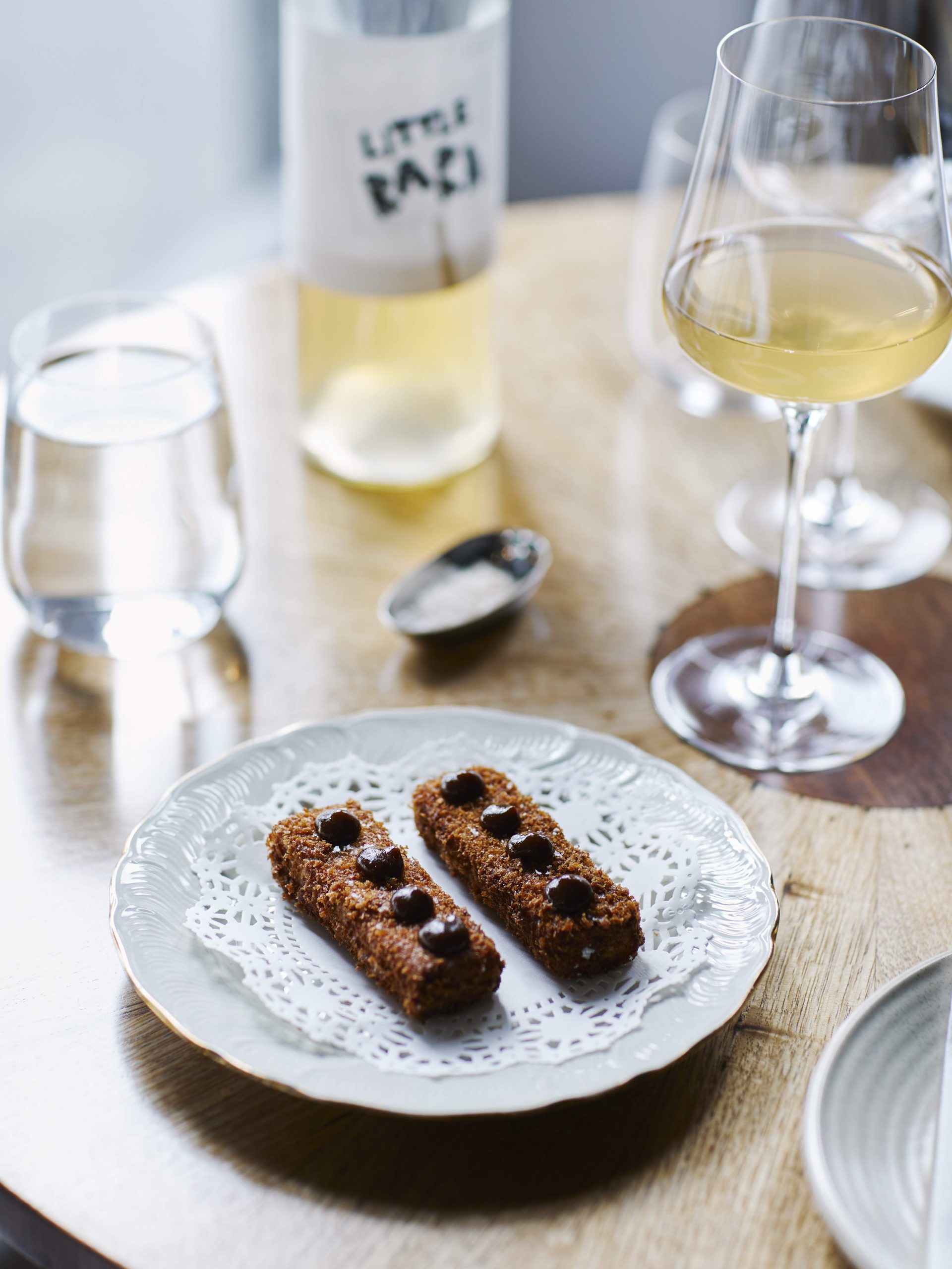 Duck croquettes sit on a doily and plate with a gentlemen’s relish on top, accompanied by a glass of wine.