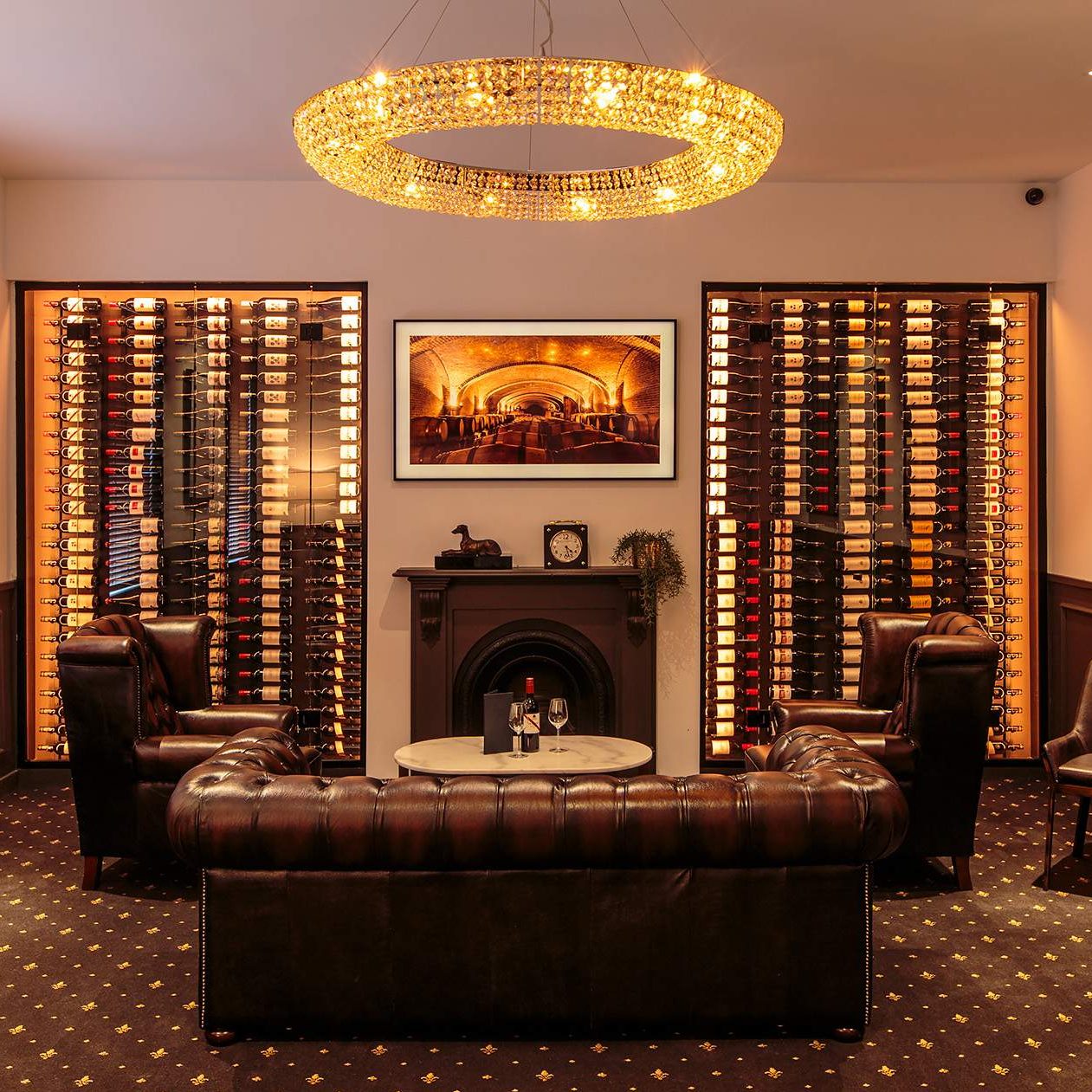 A pub lounge with a fireplace, a leather sofa and two armchairs, two wine cellars, a painting on the wall and a hanging light.