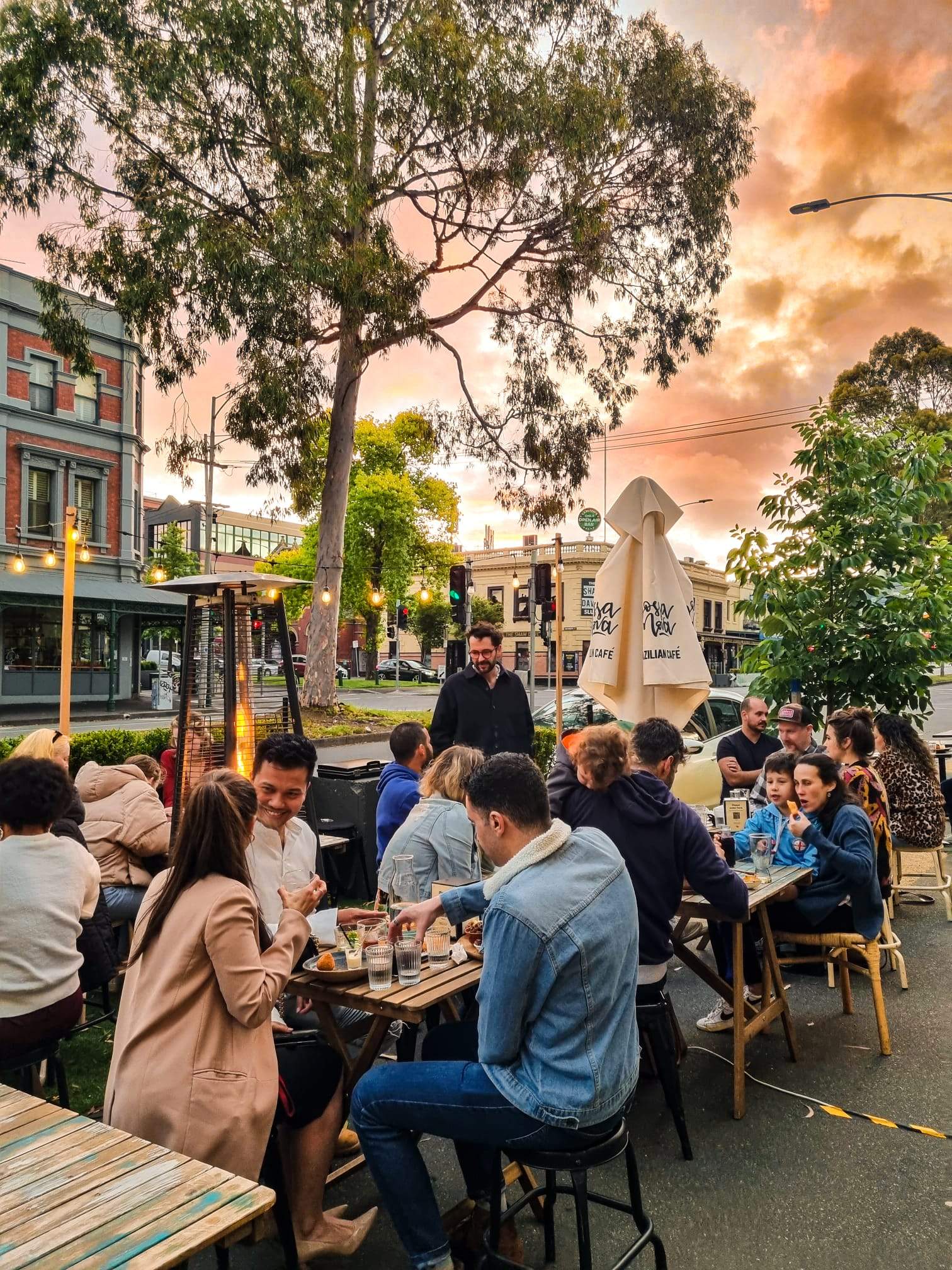 People sitting outdoors on a footpath under a setting sun with trees and buildings in the background.