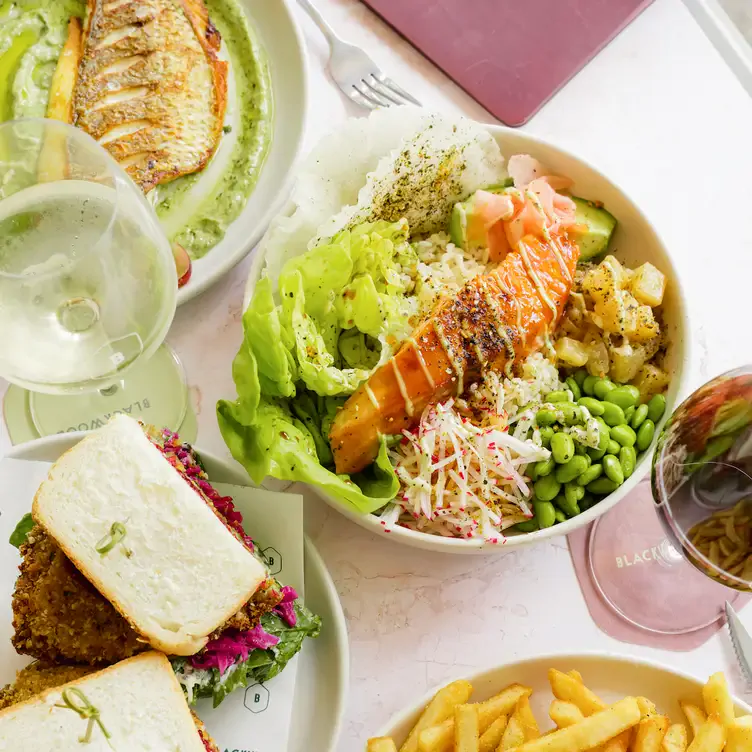 A bowl of salmon poke bowl sits next to toasts, fries, fish, salad and wine at Blackwood - Bondi in Sydney