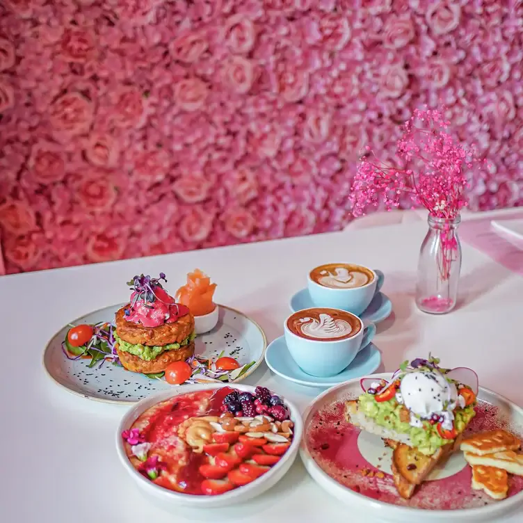 Three brunch plates sit next to two cup of coffees on a white table with a pink flower-filled background at Feekah, a restaurant in Melbourne