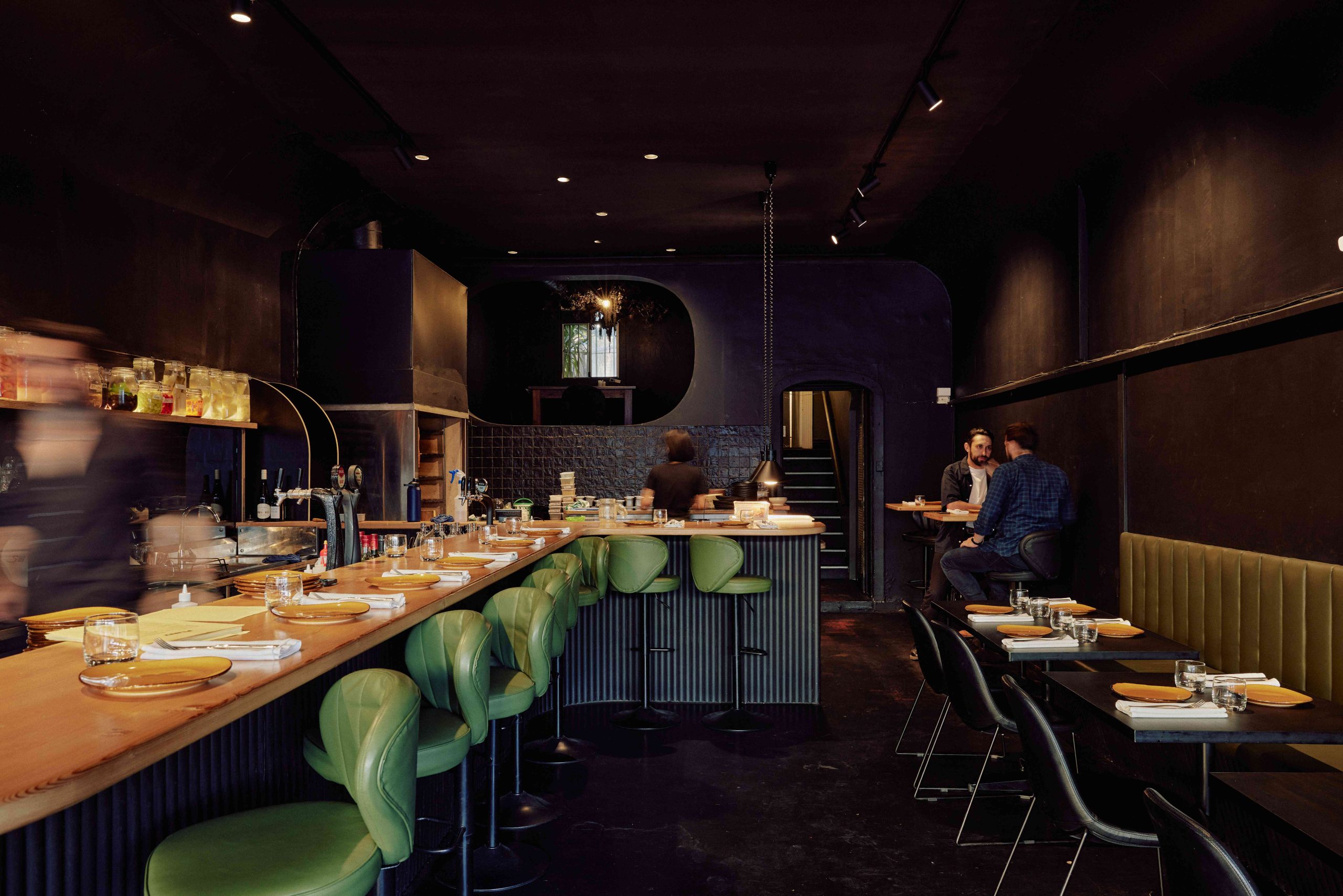 A long wooden bar, open kitchen, green bar stools, and black tables and chairs make up the dining room at FLINT in Melbourne.