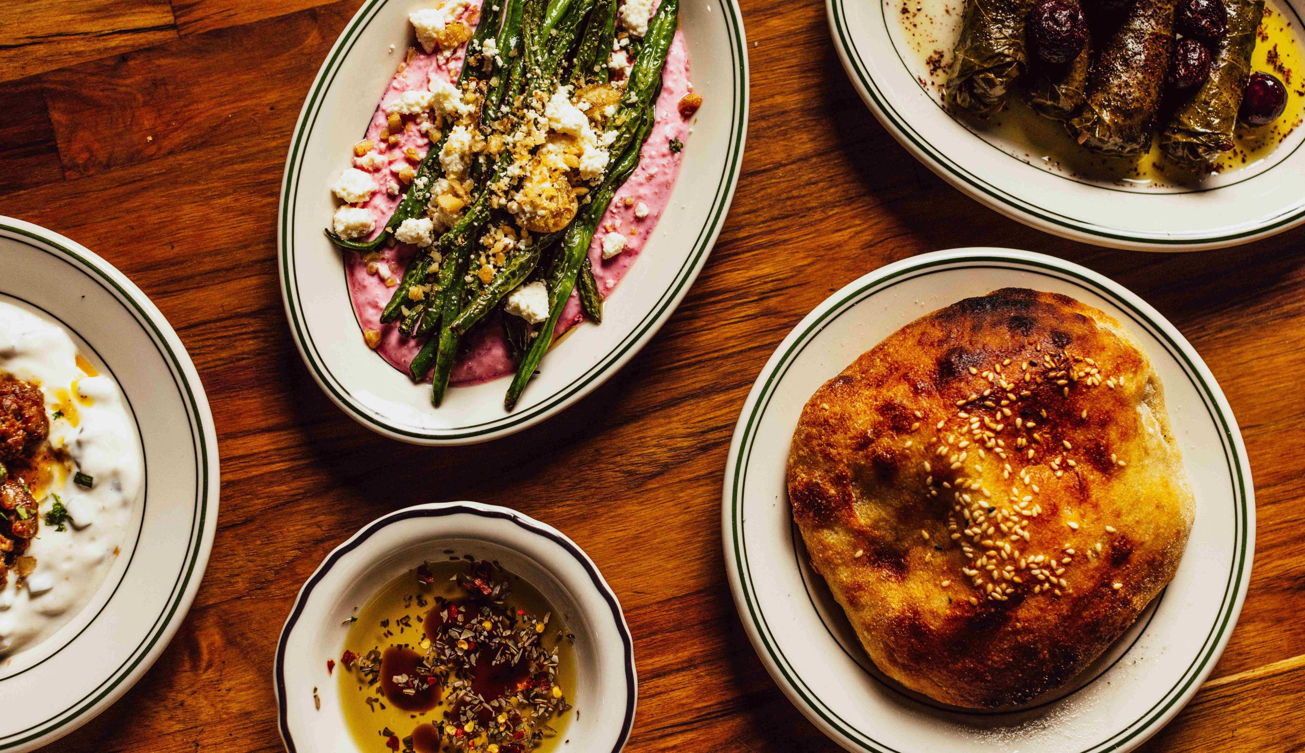 A selection of mezze including puffy bread served with chilli olive oil, stuffed vine leaves, and garlic green beans on beetroot yoghurt sitting atop a wooden table at Yakamoz Mediterranean restaurant in Melbourne.