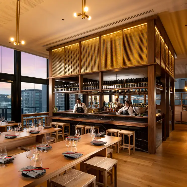 The interior of Koomo Restaurant and Bar with a few long wooden tables and a large central bar where bartenders prepare drinks.