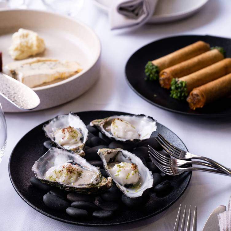 Four oysters sit on a black plate alongside other smal dishes at eleven Restaurant & Piazza in Adelaide