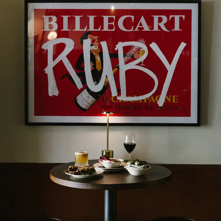 A round table with dishes, a cocktail and a glass of red wine in front of a large print with ‘Ruby’ painted over it at Ruby Dining.