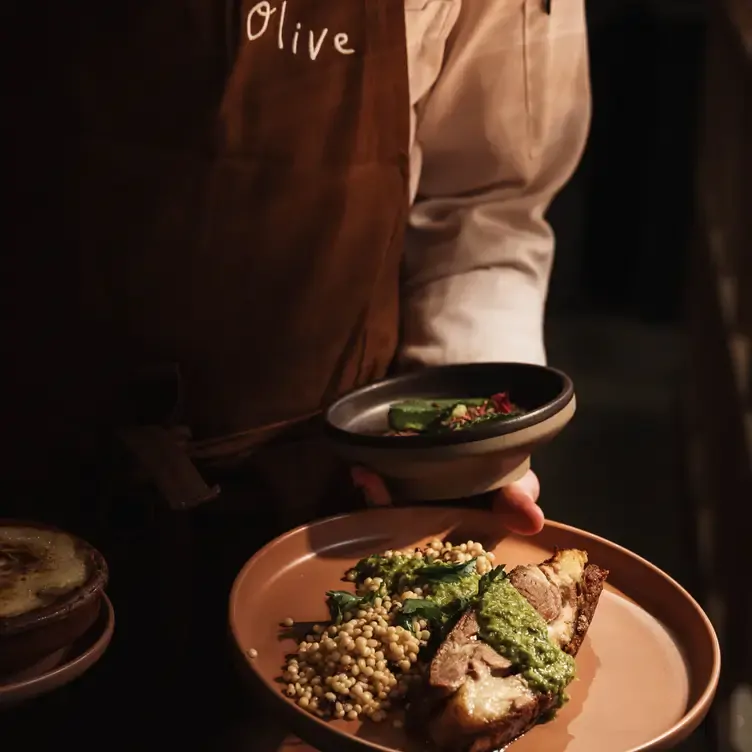 A staff member at Olive carries multiple dishes including a meat dish topped with a green sauce and with a grain-based salad on the side.