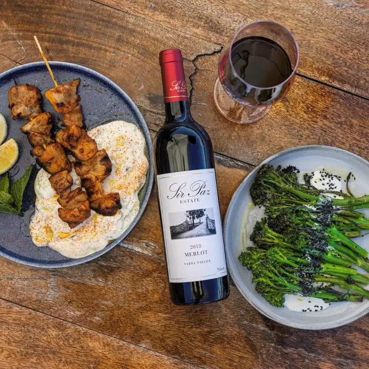 A plate of chicken skewers and a plate of broccoli served alongside a bottle of Sir Paz Estate Merlot.