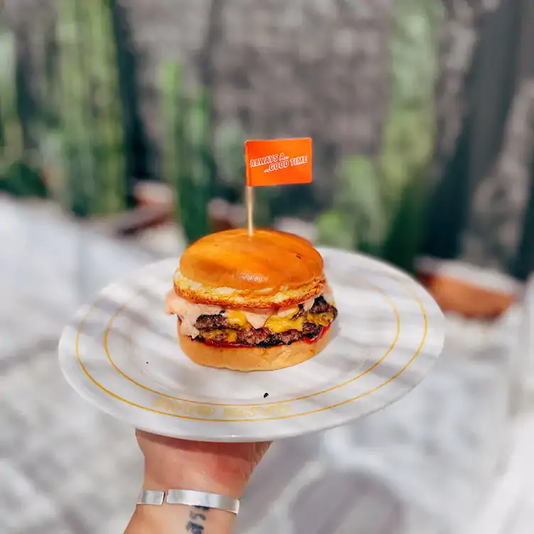 Someone holds up a burger with a small flag, in the outdoor dining space at GANG GANG Parkside, one of Adelaide’s best brunches.