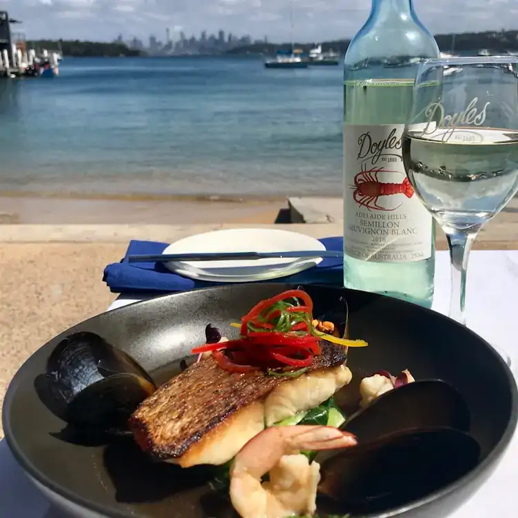 A seafood dish served alongside white wine at an outdoor table with a view of the beach and the bay at Doyles on the Beach, one of the best seafood restaurants in Sydney.