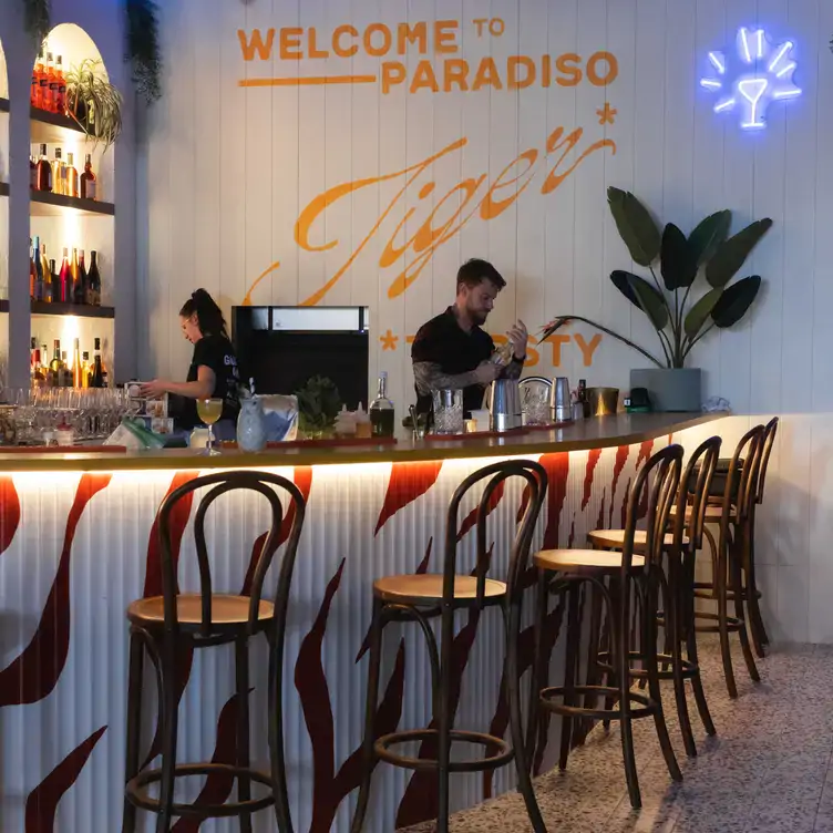 The tiger-striped bar at 99 Gang Social with a small neon-lit martini glass and a painted sign reading ‘Welcome to Paradiso, Tiger’ on the wall.