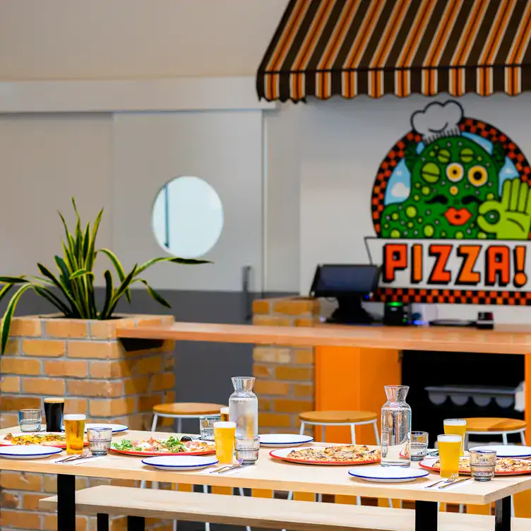 A long table and benches with pizzas and beers in front of colourful artwork at KAIJU! Beer & Pizza, one of the best family restaurants in Melbourne.