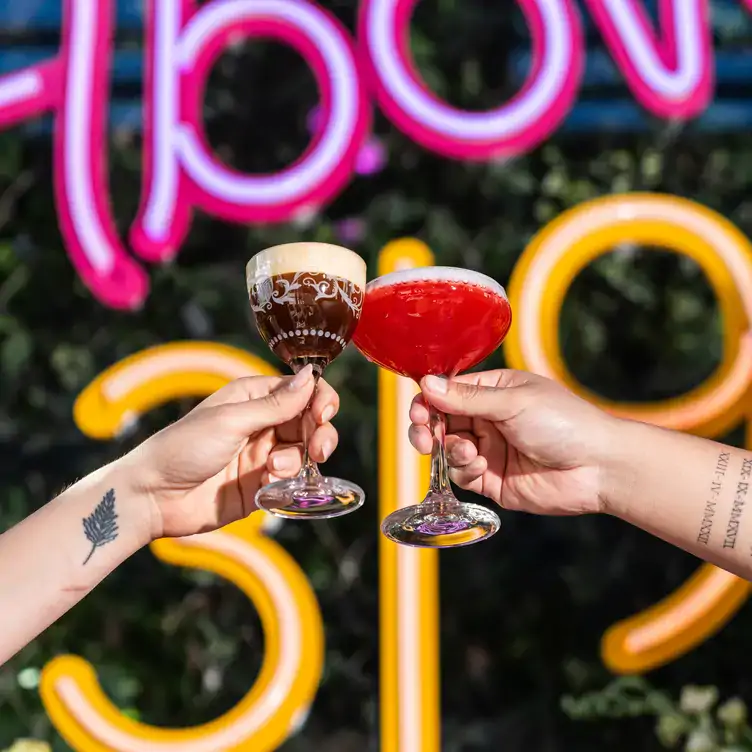 Two diners clink a brown cocktail and a red cocktail in front of a neon sign at Above 319, one of the best cocktail bars in Sydney.