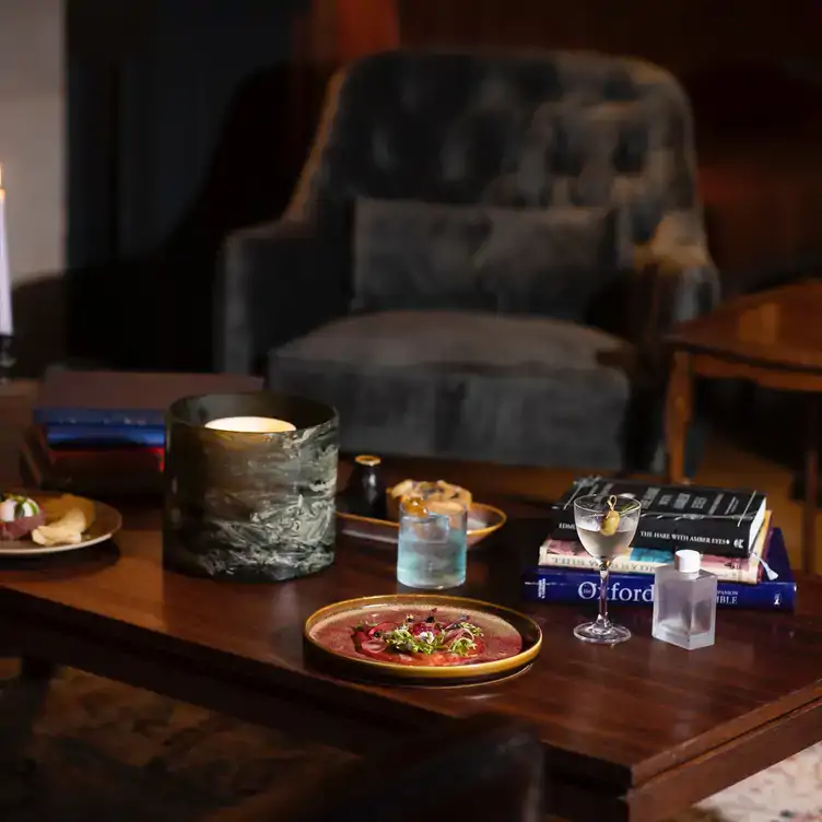 A black velvet armchair in front of a coffee table with a candle, books, cocktails and dishes served at Frank Mac’s, one of Sydney’s best cocktail bars.