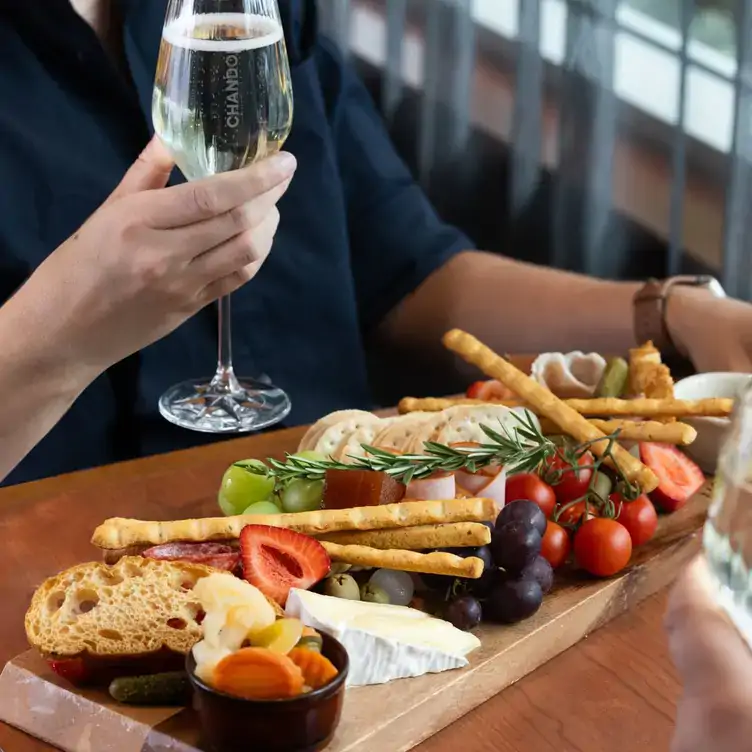 A diner holds a glass of wine while sitting at a table with a charcuterie board with fruit, cheese and bread at Sen5es Restaurant and Wine Bar.