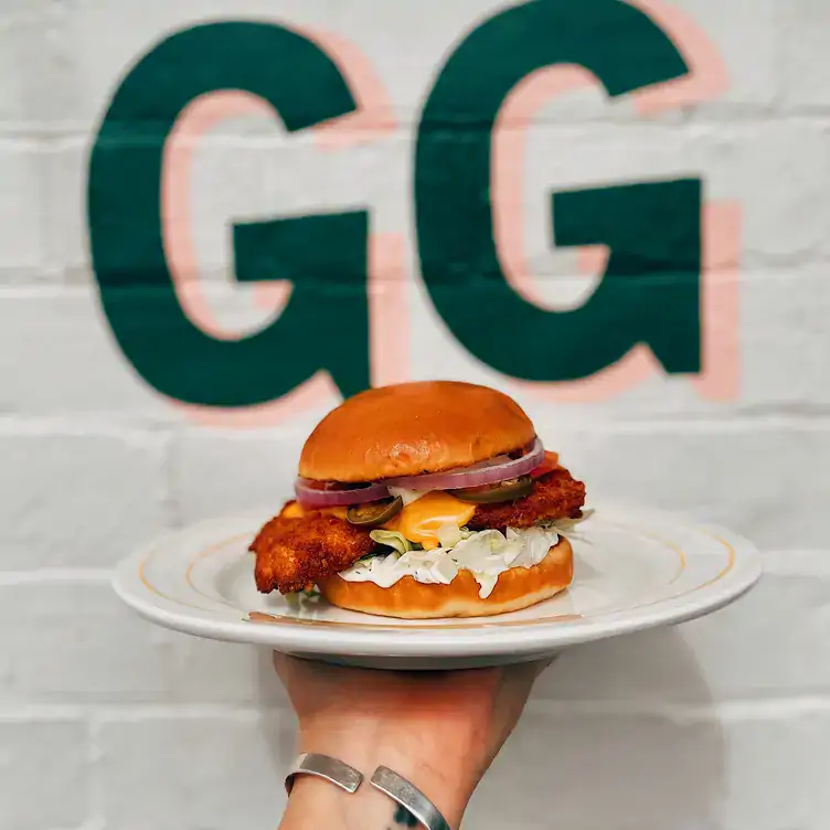 Someone holds a plate with a chicken burger in front of a green and pink ‘GG’ sign at GANG GANG Parkside.