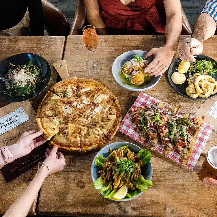 Diners reach across a table with multiple dishes including pizza, arancini balls and calamari served at Pacers & Chasers, one of the best pubs in Brisbane.