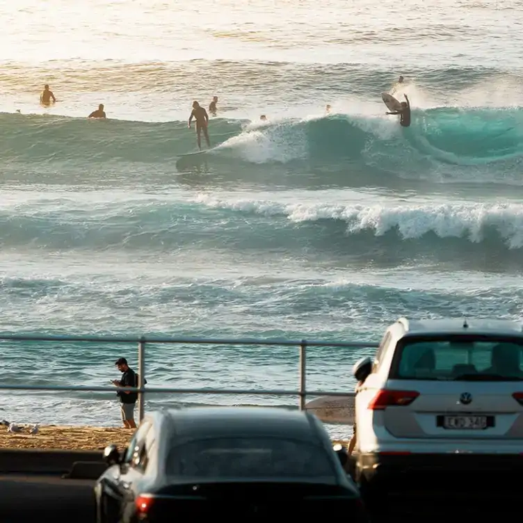 A view over the car park and towards the ocean at Bondi Beach where multiple surfers ride the waves.