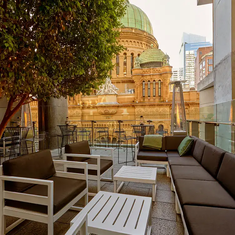 Brown-cushioned benches and chairs on a rooftop with a tree opposite the Queen Victoria Building at Zeta Bar, one of the best rooftops in Sydney.