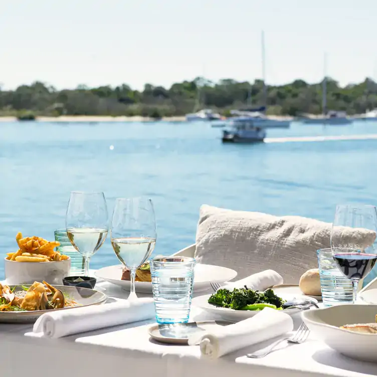 An outdoor table with salad, chips and wine in front of the Noosa River with boats on the water at Rickys River Bar, one of the best restaurants on the Sunshine Coast.﻿